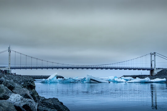 View of glacial icebergs floating serenely in the lagoon's dark waters, reflecting the sky, with a bridge spanning the distance, Glacier Lagoon, Sveitarfelagid Hornafjordur, Iceland.