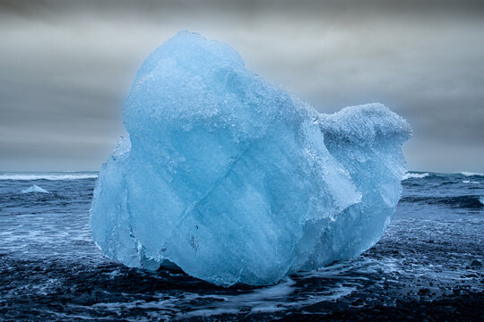 View of crystalline blue iceberg rests on the black volcanic sand beach, contrasting against the turbulent, dark waves and brooding sky, Jokulsarlon, Sveitarfelagid Hornafjordur, Iceland.