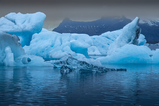 View of glacial icebergs, tinted in shades of blue, float serenely on the still waters beneath a misty sky, Glacier Lagoon, Sveitarfelagid Hornafjordur, Iceland.
