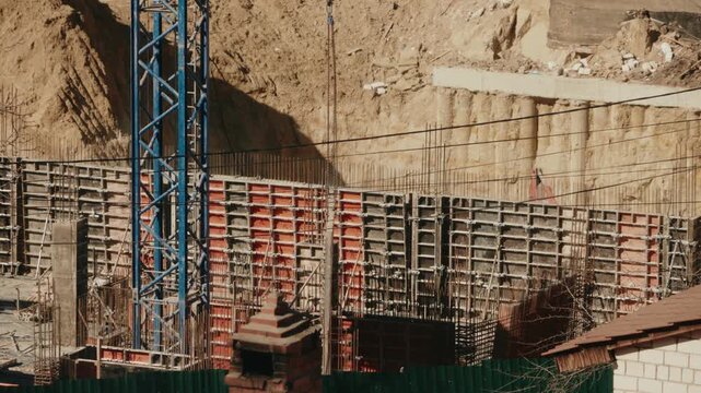 Industrial construction crane lowering large red metal formwork into a deep foundation pit for pouring reinforced concrete retaining walls at a new building site on a sunny day