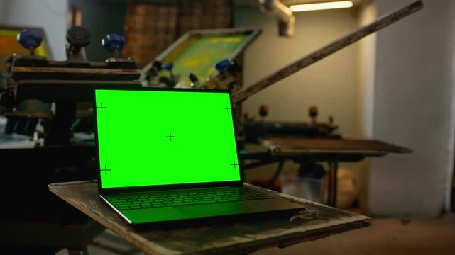 Close focus on laptop with green screen while screen printing machine rotatng with frames in workshop background