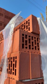 Close-up red bricks stacked outdoors, plastic wrap glistening in sunlight, perforated hollow bricks on pallet at hardware store