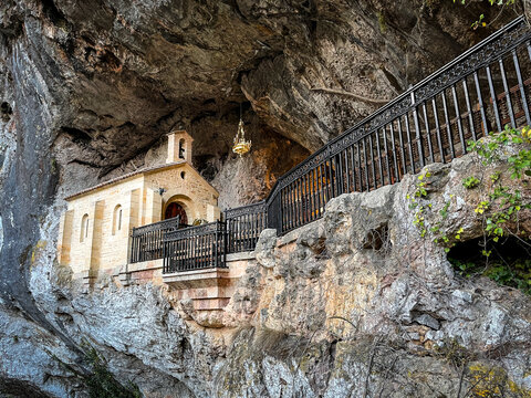 View of a small, light chapel clinging to a dark, rugged cave wall, contrasted by an elegant black railing, Santa Cueva de Covadonga, Cangas de Onis, Spain.