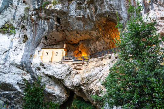 View of a light building within a vast, dark cave, warmly illuminated, surrounded by vibrant green foliage, Santa Cueva de Covadonga, Cangas de Onis, Spain.