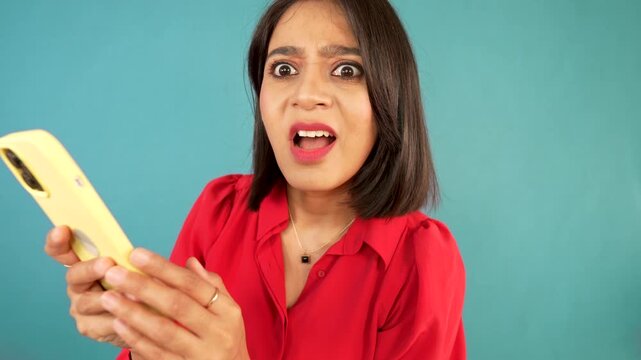 A shocked South Asian model reacting with wide eyes and open mouth while holding a yellow phone, against a bright blue background.