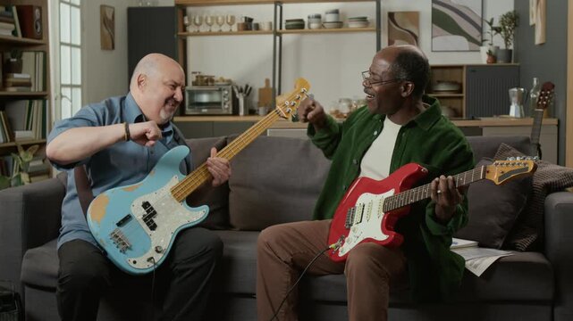 Front view shot of two senior friends playing music together sitting on couch at home and high five in successful jam session, active hobby in retirement, senior friendship
