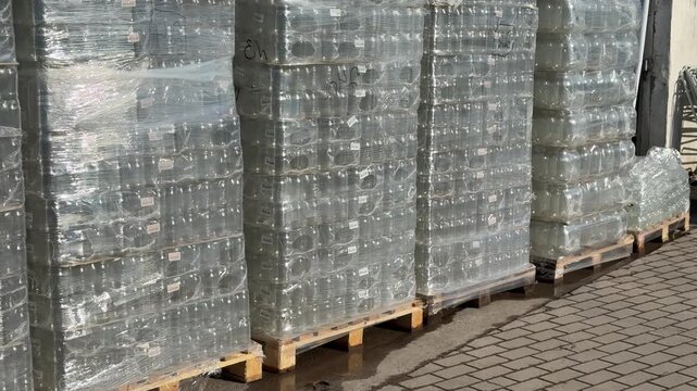 Pallets of glass jars wrapped outdoors, wooden pallets lined on sidewalk outside hardware store, shrink-wrap glinting in sunlight