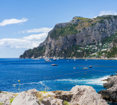 View of the turquoise sea reflecting the azure sky, speckled with boats and framed by rugged cliffs and white rocks, Capri, Campania, Italy.
