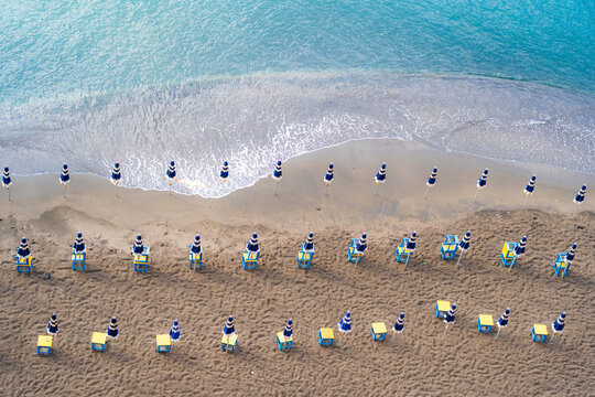 View of orderly rows of vibrant blue and yellow beach umbrellas and chairs line the sandy shore where the turquoise sea gently meets the land, Capri, Campania, Italy.