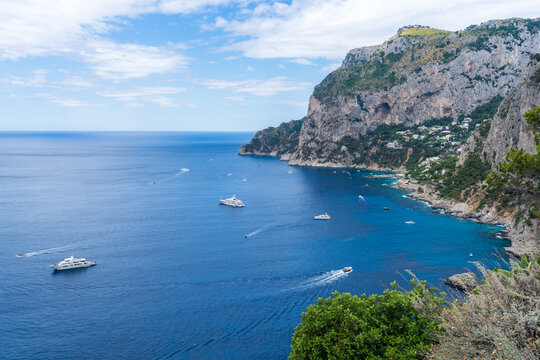 View of deep blue sea dotted with boats contrasting against the craggy green cliffs under a bright sky, creating a serene coastal panorama, Capri, Campania, Italy.