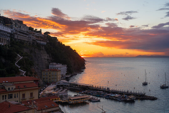 View of the sun ablaze over the sea, casting a warm glow on the harbor's boats and the cliffside buildings, a serene Italian vista, Sorrento, Campania, Italy.