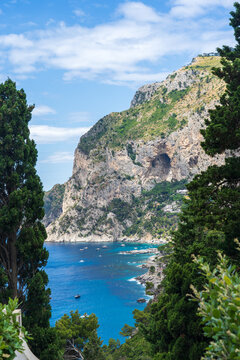 View of turquoise waters meet rugged cliffs, framed by lush greenery, creating a serene coastal vista under a bright sky, Capri, Campania, Italy.