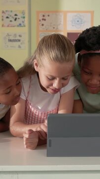 Vertical video: Leaning in center girl in pinafore tapping gray tablet in classroom sharing work