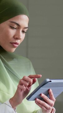Vertical video: Tapping focused woman in green hijab, scrolling tablet at home, reading with cup