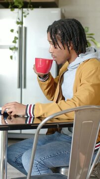 Vertical video: Working on computer, man in mustard coat sipping red mug using mouse in kitchen