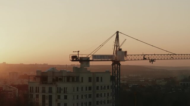 Silhouette of a large tower crane slowly rotating at a construction site against the beautiful golden hour sky, representing urban development, industrial machinery, and real estate growth. timelapse