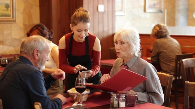 Positive female server listening carefully while standing beside old couple enjoying evening meal in restaurant