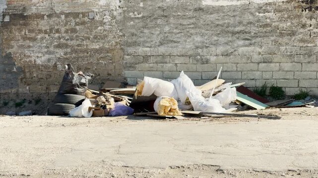 A messy pile of garbage, tires, and white sacks sits against a weathered wall. The bright sunlight exposes the neglected state of the lot. This scene feels forgotten, messy, and lonely.