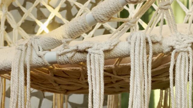 Intricate cotton knots secure a bohemian swing chair. The soft fringe drapes elegantly, evoking a sense of calm and relaxation. It sits nestled in a bright, tranquil outdoor space.