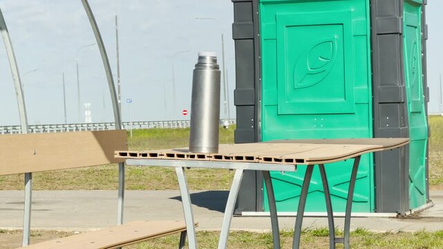 A silver thermos rests on a wooden table, providing a refreshing break for travelers. The green public toilet stands nearby under a bright sky. This scene evokes a sense of calm and relief.
