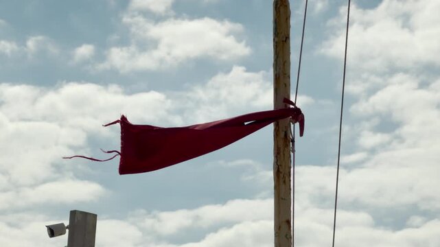 A worn red flag flaps wildly on a weathered pole against a bright, cloudy sky. The frayed fabric evokes a sense of grit and endurance. It feels lonely yet brave in the strong wind.