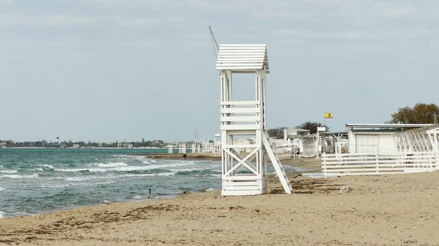 A tall white wooden tower stands on the sandy beach as waves crash nearby. The scene feels lonely yet peaceful under the wide, pale sky. It captures a quiet moment of coastal beauty.