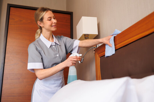 Hotel housekeeper wiping wooden bed headboard with microfiber cloth and cleaner