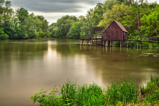 View of a rustic watermill house standing on stilts over the tranquil, glassy lake reflecting the cloudy sky and lush green trees, Tomasikovo, Slovakia.