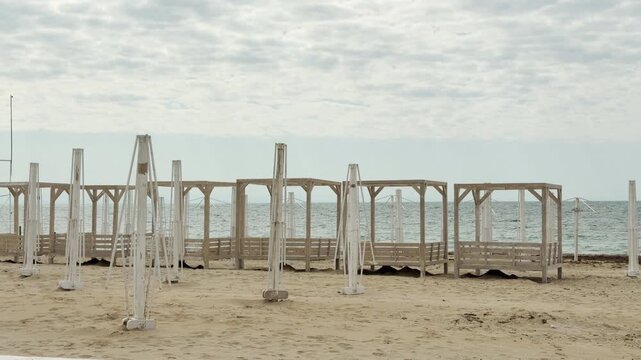 Empty wooden gazebos and folded white umbrellas sit on a vast, sandy beach. The overcast sky and calm sea create a lonely, nostalgic feeling of a quiet off-season. It is a peaceful, still scene.