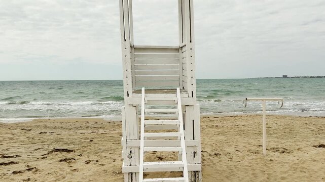 A tall white lifeguard tower stands firmly on the empty sand. The vast sea and cloudy sky create a lonely, protective atmosphere. It feels like a quiet guardian during the cold off-season.