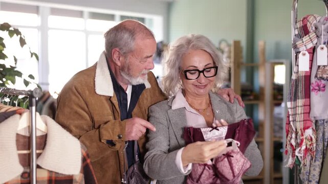 Mature husband and wife stand in a clothing boutique and choose underwear. Elderly couple buying a bra set in a lingerie store. High quality 4k footage