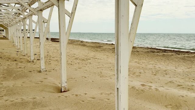 A rhythmic row of white wooden pillars supports a long shade structure on the sand. The perspective creates a lonely, nostalgic feeling of a quiet resort before the summer crowds.