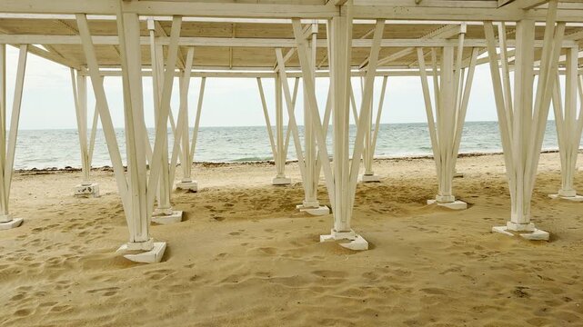 Sturdy white wooden pillars hold up a large canopy over the soft sand. The perspective creates a sense of calm and geometric beauty. This quiet scene feels peaceful, steady, and very relaxing.