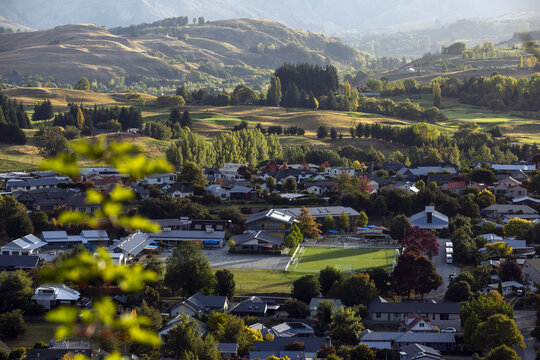 View of late afternoon sunlight across the town nestled amongst rolling hills, with a vibrant green fields contrasting against the roofs, Arrowtown, Otago Region, New Zealand.