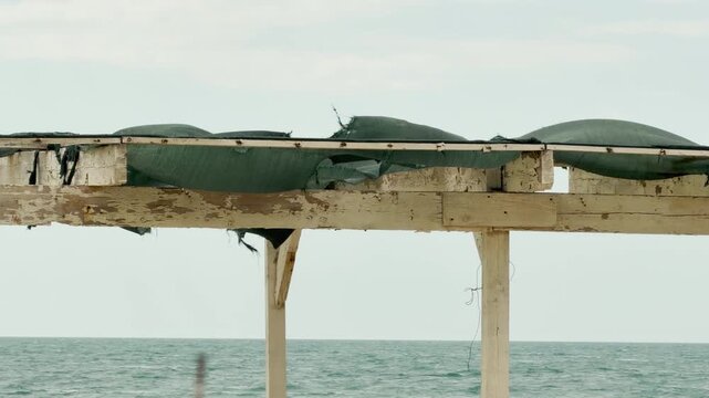 A torn green net sags from an old wooden frame by the sea. The peeling paint and gray water create a sad, lonely mood. It looks like a quiet place forgotten after a busy summer.