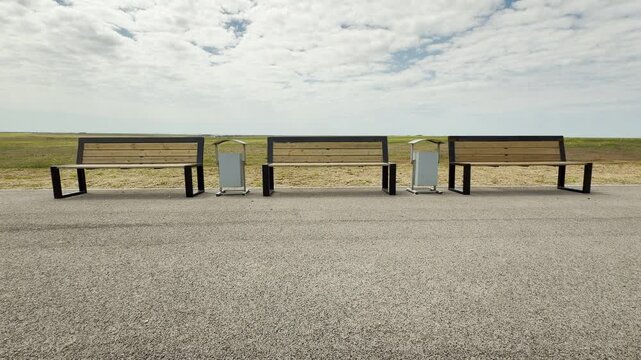 A row of modern wooden benches sits lonely along a quiet roadside. The vast open field and heavy clouds create a peaceful yet melancholy mood. This still scene feels calm and solitary.