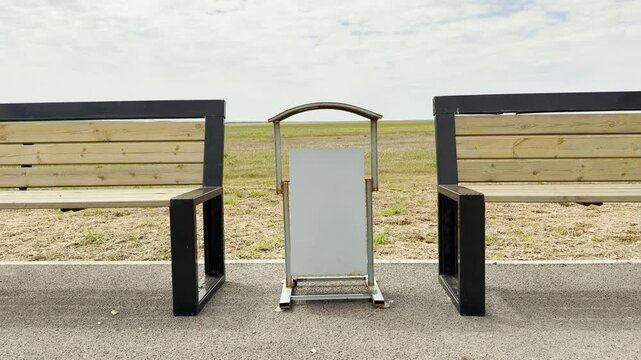 Two wooden benches sit quietly beside a small bin in a lonely park. The wide field and cloudy sky create a peaceful, solitary mood. This simple scene feels calm and still.