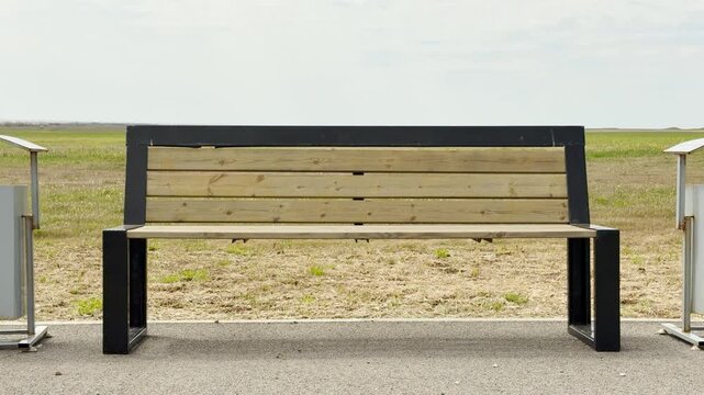 A clean wooden bench sits quietly between two metal bins in a peaceful park. The vast green field behind it creates a sense of calm and solitude. This still scene feels fresh and inviting.