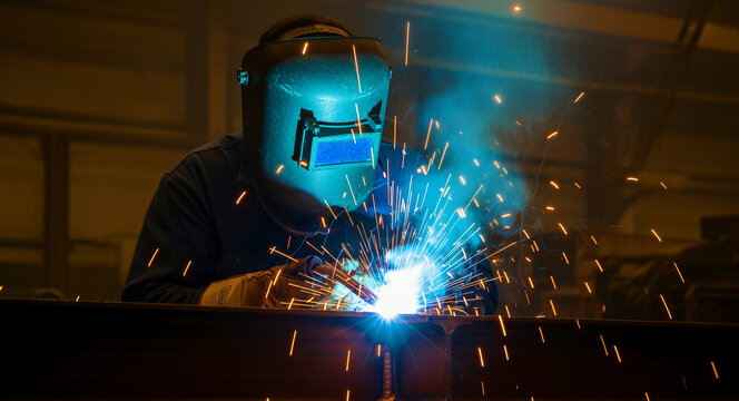 Welder performing arc welding on a steel beam in a workshop. Intense sparks and bright light illuminate the worker in a protective mask. Industrial manufacturing concept for engineering services.