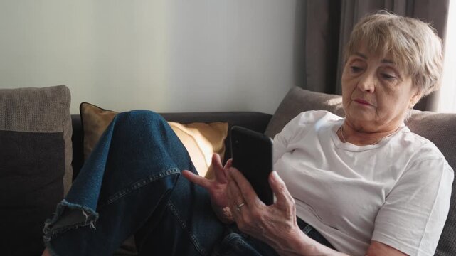 Senior woman relaxing on a sofa in a cozy living room, using smartphone. Authentic everyday moment, modern lifestyle, technology adoption by elderly at home.