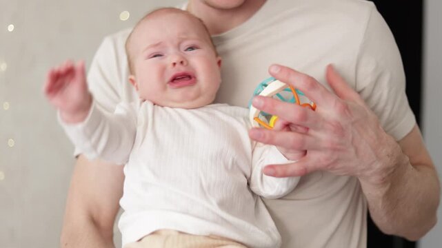 Father comforting crying baby with a colorful rattle toy