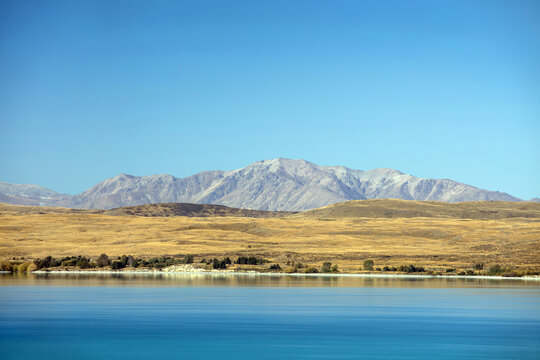 View of the brilliant turquoise waters reflecting a golden-brown landscape leading to rugged grey mountains under a vast blue sky, Ben Ohau, Canterbury Region, New Zealand.