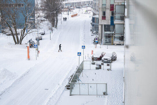 View of a solitary figure braving the snow-covered streets, with modern buildings and parked cars under a blanket of white, Tallinn, Estonia.