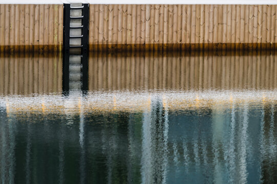 View of the tranquil waters mirroring the adjacent wooden structure and metallic ladder, blurring the line between reality and reflection, Tallinn, Estonia.