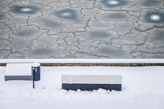 View of a snow-covered bench and bin stand in stark contrast to the cracked ice patterns on the frozen body of water, Tallinn, Estonia.