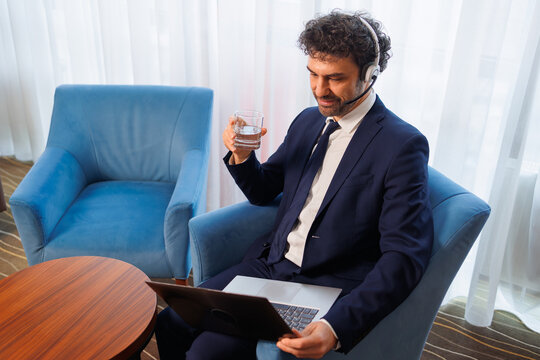 Smiling Businessman in Suit with Headset Using Laptop and Holding Glass of Water During Video Call