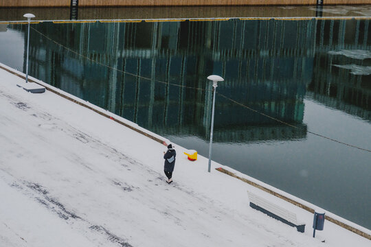 View of a solitary figure trekking through the snow-dusted ground along the water's edge, with mirrored buildings reflecting in the dark surface, Tallinn, Estonia.