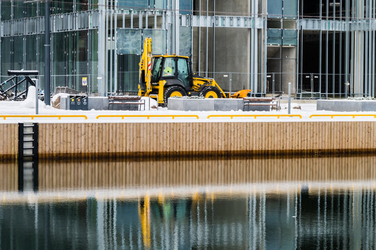 View of a yellow digger stands poised near the waterfront, its reflection shimmering on the dark water against the backdrop of a modern building, Tallinn, Estonia.