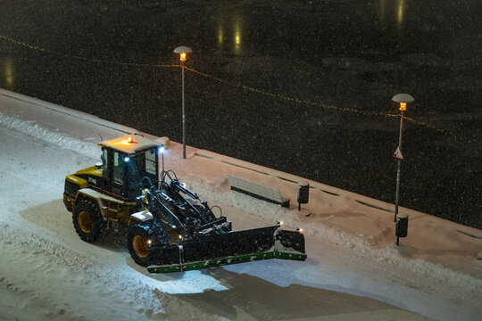 View of a yellow snowplow carving a path through fresh snow under the faint glow of streetlights along a dark, wintry waterfront, Tallinn, Estonia.
