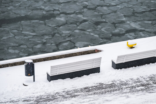 View of icy waters contrast with the snowy banks, benches, and a yellow bollard in a serene yet chilly winter scene, Tallinn, Estonia.